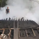 Mężczyzna nadzoruje proces, jak powstaje węgiel drzewny. Dym unosi się z kopca, ukazując tradycyjną metodę produkcji.