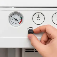 Dłoń reguluje pokrętło, kontrolując dopuszczanie wody do pieca. Manometr wskazuje ciśnienie.