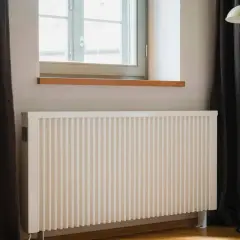 Biały kaloryfer pod oknem, obok ciemnej zasłony i lampy stojącej. Dowiedz się, jak ustawić kaloryfery przy ogrzewaniu gazowym dla optymalnego komfortu.