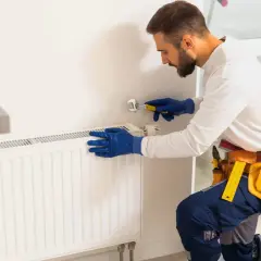 Mężczyzna w niebieskich rękawiczkach używa klucza do odpowietrzenia grzejnika aluminiowego.
