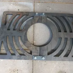 Metalowy ruszt awaryjny do pieca na ekogroszek, z wycięciami i śrubami, leży na betonowych płytkach.