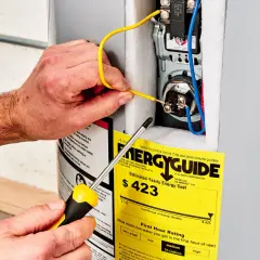 Ręce majsterkowicza podłączają przewody do bojlera, obok żółtej etykiety EnergyGuide, która informuje, ile prądu pobiera grzałka do bojlera.