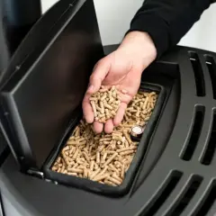 Dłoń wsypuje pellet do zasobnika pieca. Czy piec na pellet musi pracować cały czas? Odpowiedź zależy od modelu i ustawień.