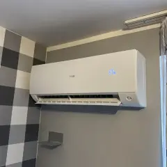 Biała klimatyzacja Haier zamontowana na szarej ścianie, obok wzorzystej tapety i zasłon.