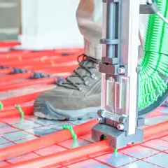 Instalator z narzędziem układa rury do ogrzewania podłogowego. Czerwone rurki na zielonych klipsach tworzą siatkę.