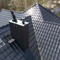 Nowoczesny, czarny komin do pieca na ekogroszek na błyszczącym dachu z dachówki. Obok okno dachowe.