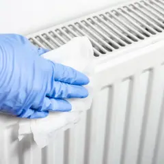 Rękawiczka w niebieskiej rękawiczce czyści biały grzejnik aluminiowy wilgotną ściereczką, pokazując, jak wyczyścić grzejnik aluminiowy w środku.