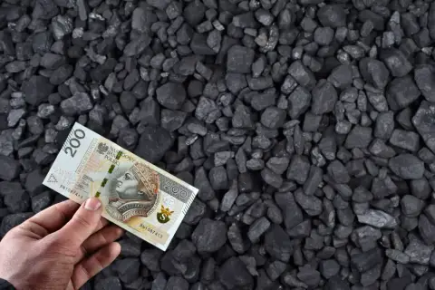 Dłoń trzyma banknot 200 zł na tle węgla. Zastanawiasz się, ile kosztuje węgiel tona?