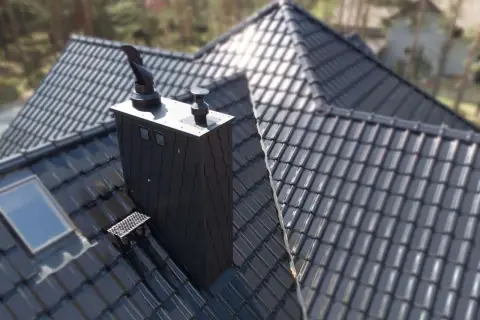 Nowoczesny, czarny komin do pieca na ekogroszek na błyszczącym dachu z dachówki. Obok okno dachowe.