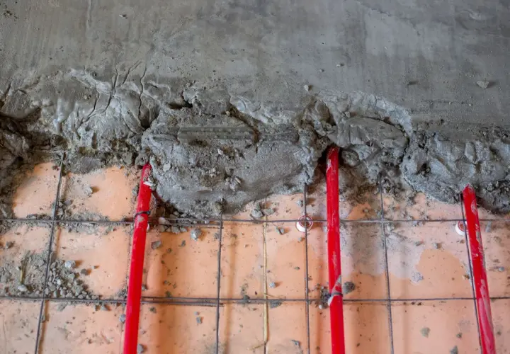 Czerwone rury ogrzewania podłogowego pod świeżą wylewką betonową. Kluczowe jest odpowiednie dobranie grubości wylewki, by zapewnić efektywne rozprowadzanie ciepła.