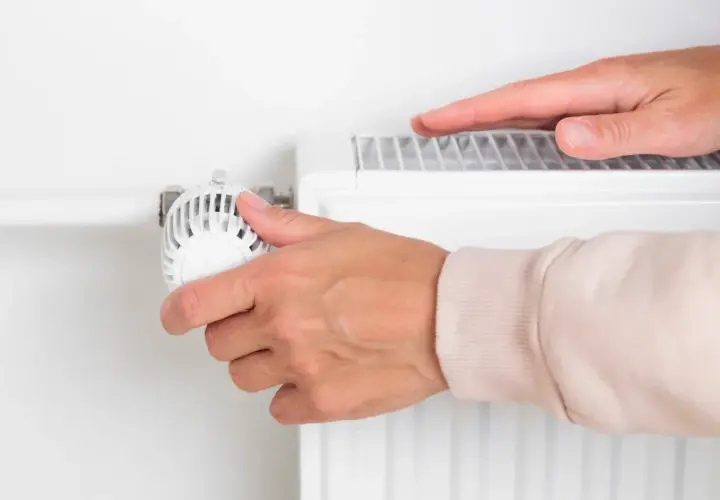 Dłoń reguluje termostat grzejnika, zastanawiając się, kiedy wyłączyć ogrzewanie w domu, by oszczędzić energię.
