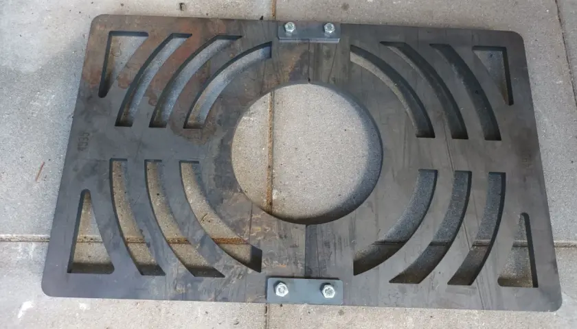 Metalowy ruszt awaryjny do pieca na ekogroszek, z wycięciami i śrubami, leży na betonowych płytkach.