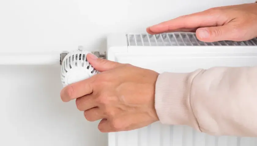 Dłoń reguluje termostat grzejnika, zastanawiając się, kiedy wyłączyć ogrzewanie w domu, by oszczędzić energię.