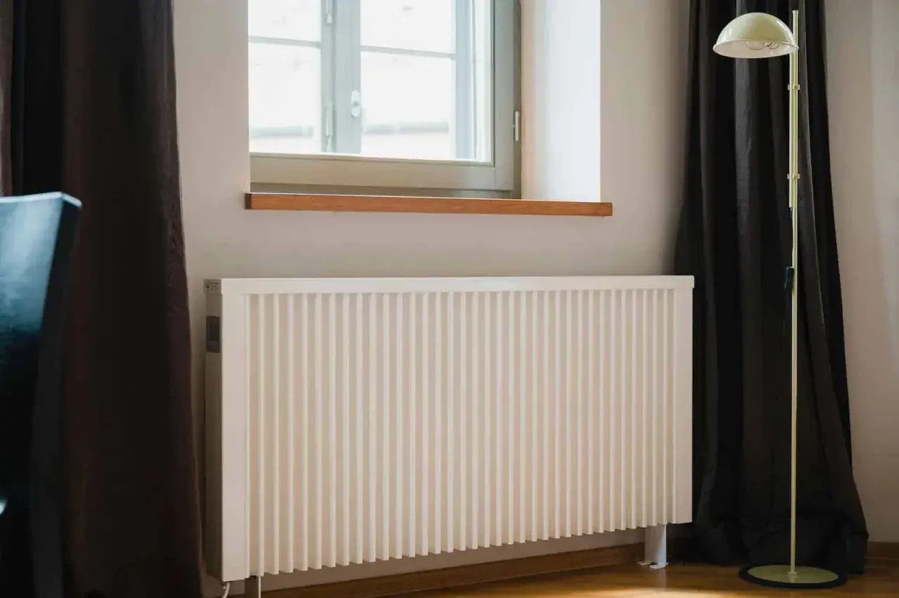 Biały kaloryfer pod oknem, obok ciemnej zasłony i lampy stojącej. Dowiedz się, jak ustawić kaloryfery przy ogrzewaniu gazowym dla optymalnego komfortu.