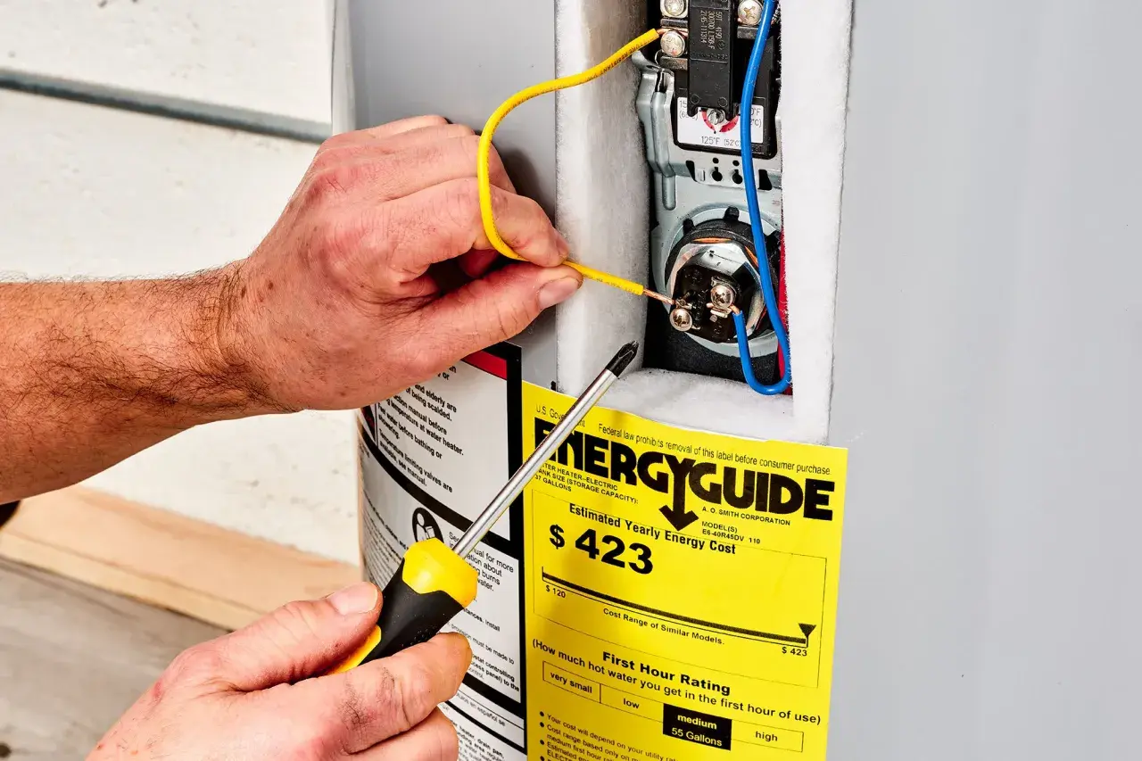 Ręce majsterkowicza podłączają przewody do bojlera, obok żółtej etykiety EnergyGuide, która informuje, ile prądu pobiera grzałka do bojlera.