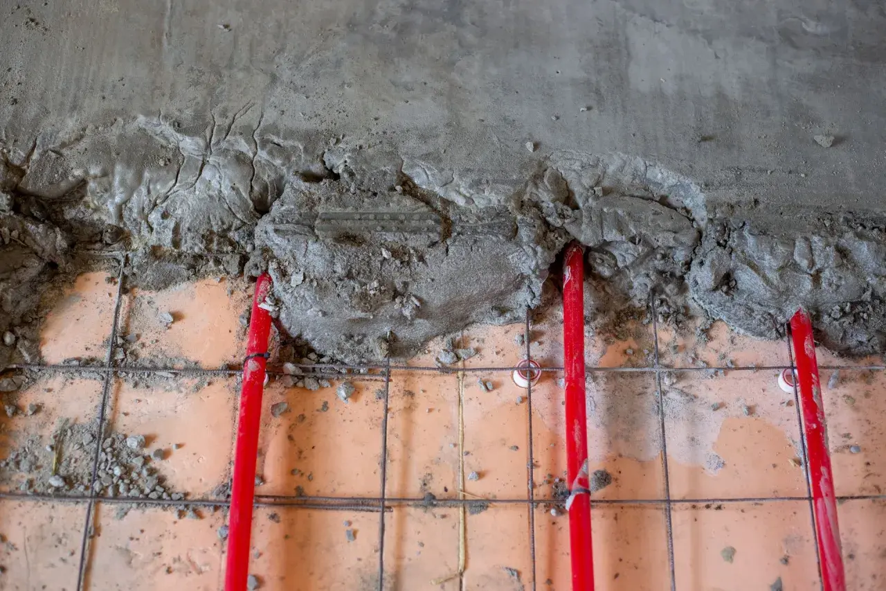 Czerwone rury ogrzewania podłogowego pod świeżą wylewką betonową. Kluczowe jest odpowiednie dobranie grubości wylewki, by zapewnić efektywne rozprowadzanie ciepła.