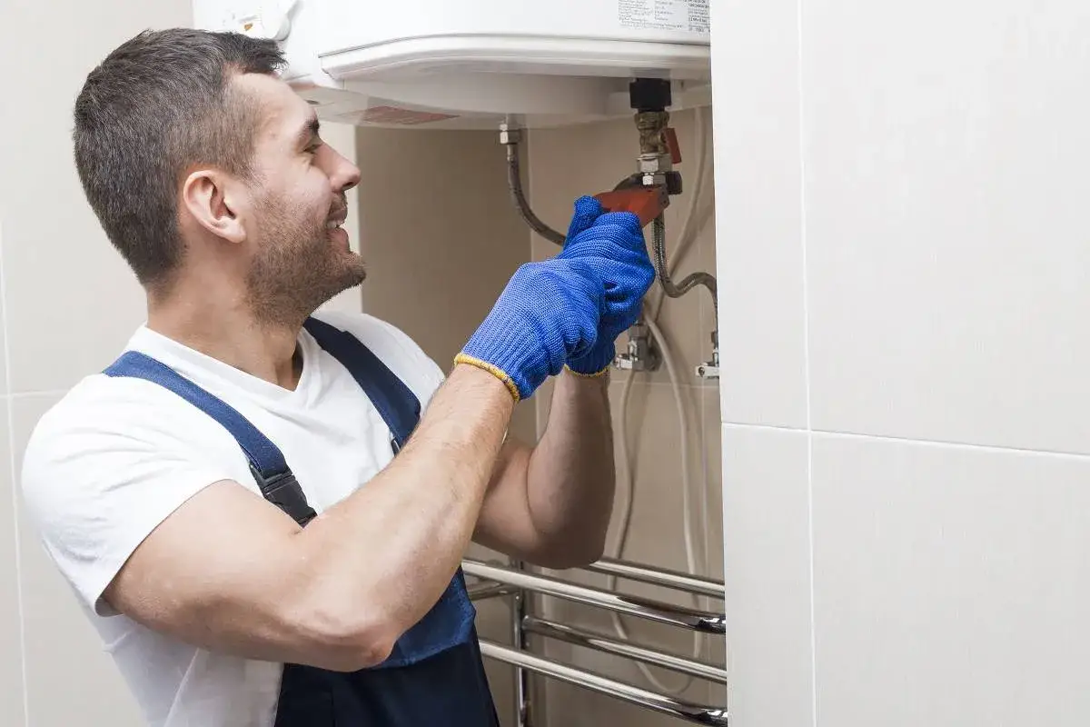 Instalator w niebieskich rękawicach próbuje spuścić wodę z bojlera bez zaworu spustowego, używając klucza.